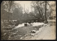 Lightly sepia-toned photograph showing a wooden dam in Bassett Creek. Around the creek and dam are dirt paths, bare trees and wooden houses in the background.